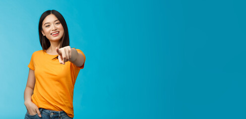 Wall Mural - Lucky winner. Cheerful charismatic asian cute urban girl stand yellow t-shirt smiling friendly pointing finger camera choosing, picking person, inviting you team, stand happy blue background