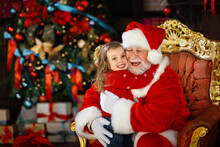 Santa Claus Hugs The Child And Promises To Fulfill His Cherished Wish. A Smiling Little Girl On Santa's Lap.