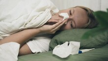 A sick girl lies on the bed covered with a blanket, blowing her nose into paper napkins. The concept of medicine and health.