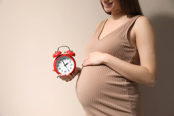 Poster - Young pregnant woman holding alarm clock near her belly on beige background, closeup and space for text. Time to give birth