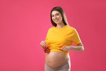 Wall Mural - Young pregnant woman holding alarm clock near her belly on pink background. Time to give birth