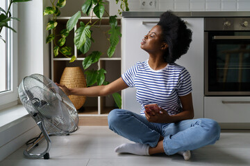 Young black woman sits on floor with smartphone, tries to catch air currents emanating from electric fan. African American lady spends time in hot apartment and wants to cool off playing mobile apps