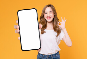 Cheerful beautiful Asian woman holding smartphone and shows ok sign on light yellow background.