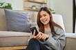 © bongkarn - Happy young Asian female sits in her living room and uses her smartphone