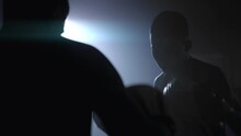 Two Young Male Boxers Sparring, Combat Sports Training. A Couple Of Fighters Practising In The Dark Gym, One Is Seen From The Back Wearing Boxing Gloves, Punching, Second Is Facing Him In Boxing Pads