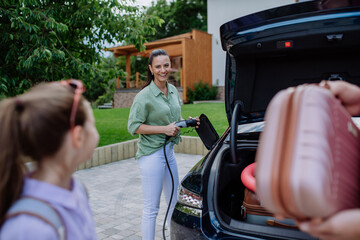 Happy mother holding power supply cable and charging their electric car, rest of family putting suitcases in car trunk, preparating for holidays.