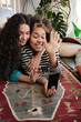 © Clique Images - Young lesbian couple spending time at home lying on floor greeting their friend on video call using smartphone