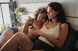 © Clique Images - Young Black and Caucasian women in love relaxing together on bed in morning surfing Internet on smartphone