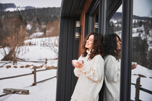 Curly Young Woman Resting On Terrace Of Modern Barn House In The Mountains. Portrait Of Happy Female Tourist Holding Cup Of Tea, Enjoying In New Cottage In Winter.