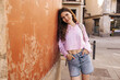 Cute young caucasian girl looks at camera leaned on wall stands at street with hands in pockets. Brown hair woman wears shirt, shorts and backpack. Concept city life.