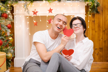 Couple in love, man and woman in white t-shirts, holding wooden red hearts, sitting on floor against the background of an artificial fireplace decorated for Christmas in the living room of the house.