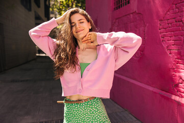 Peaceful young caucasian woman relaxing with closed eyes outside near pink brick wall in background. Brunette wears singlet, cardigan and skirt. Concept tenderness, lifestyle.