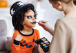 © JenkoAtaman - Festive makeup for Halloween. Woman doing pumpkin make-up for ethnic curly boy in costume while preparing holiday