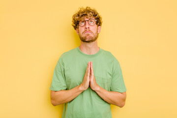 Wall Mural - Young caucasian man isolated on yellow background holding hands in pray near mouth, feels confident.