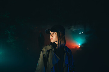 Attractive woman in stylish casual clothes stands at night in a foggy park in a colored light and looks away with a serious face.