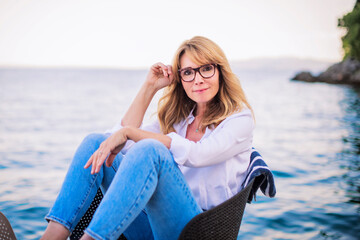 Wall Mural - Smiling mature woman wearing white shirt and blue jeans and relaxing by the sea