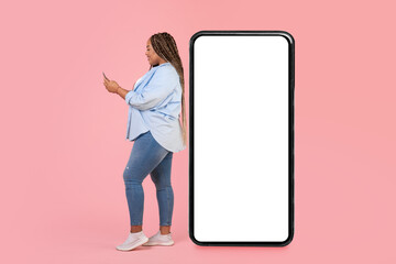 Wall Mural - Black Female Using Phone Standing Near Large Smartphone, Pink Background