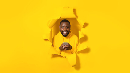 Excited african american man using mobile phone, standing breaking through yellow paper background