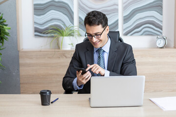 Handsome smiling businessman in black suit sitting at the desk and working on laptop in the modern office