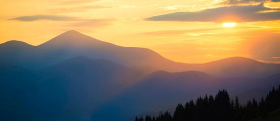Wall Mural - Sunset in mountains. Panorama mountains landscape with sun shining through orange clouds