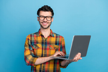 Wall Mural - Photo of cheerful positive handsome man in spectacles wear plaid shirt holding laptop online meeting isolated on blue color background