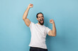 Portrait of excited man with beard wearing white T-shirt expressing winning gesture with raised fists and screaming, celebrating victory. Indoor studio shot isolated on blue background.