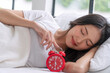 Beautiful cheerful asian woman sleeping lying down relax on pillow with blanket holding hand red alarm clock at home. Happy young girl teenager nap sleepy rest with alarm clock on white bed in bedroom