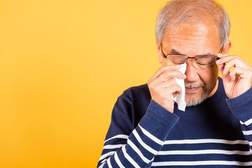 Wall Mural - Asian elder man crying raise glasses with tissue wipe red eyes studio shot isolated on yellow background, Portrait senior old man sad wiping away his tears, Upset depressed lonely