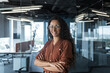 Portrait of successful young beautiful woman company owner and founder. Hispanic businesswoman standing in office wearing glasses and brown shirt, looking at camera, arms crossed, smiling.