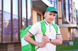 © Евгений Шемякин - portrait of young handsome happy courier guy smile, food delivery man with green thermo box for food delivering food outdoors in the yard on buildings background at summer day in cap and uniform.