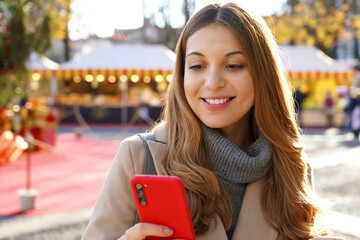 Wall Mural - Close-up of young smiling beautiful woman using smartphone on the street at winter