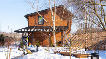 A man gardener prunes a tree with an electric chain pruner on a sunny winter day. An electric garden saw with a telescopic pole in hands of a man cutting the tree branches in garden.