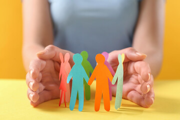 Wall Mural - Woman protecting paper human figures on yellow table, closeup. Diversity and inclusion concept