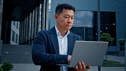 Asian 40s middle-aged businessman typing laptop outdoor in downtown Korean adult entrepreneur employer man worker manager investor architect standing near office company building working with computer