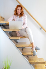 Wall Mural - Front view of ginger young woman sitting on the stairs and working on laptop