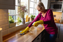 Happy Senior Caucasian Woman Wearing Rubber Gloves, Cleaning Countertop In Kitchen