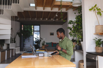 Wall Mural - Happy african american man sitting at table in kitchen using laptop and smartphone