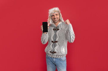 Wall Mural - glad handsome man wear sweater and winter hat hold phone, copy space, presenting product