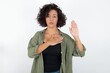 young beautiful woman with curly short hair wearing green overshirt over white wall Swearing with hand on chest and open palm, making a loyalty promise oath