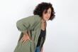 © Roquillo - young beautiful woman with curly short hair wearing green overshirt over white wall Suffering of backache, touching back with hand, muscular pain