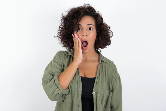 Shocked young beautiful woman with curly short hair wearing green overshirt over white wal looks with great surprisment being very stunned, astonished with unexpected news, Facial expressions concept.