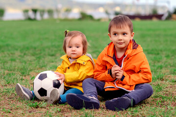 two kids, boy and toddler girl sitting on football field in summer or autumn day with soccer ball af