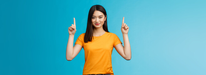 Heads up see awesome promo. Assertive good-looking confident asian woman showing friend advertisement, raise index fingers pointing top, smiling camera self-assured, stand blue background determined