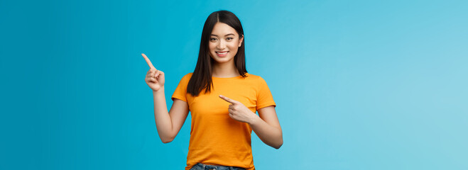 Friendly cheerful asian brunette girl inviting visit link interesting place, pointing upper right corner smiling joyfully camera, telling you about good promotion, stand blue background upbeat