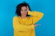© Jihan - young beautiful woman with curly short hair wearing yellow sweater over blue wall feeling positive has amazed expression, indicates something. One hand on head and pointing with other hand.