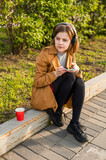 Fototapeta Boho - A young teenage girl sits on the street with a smartphone in her hand, headphones, listens to music. High quality photo