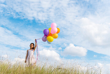 Wall Mural - Cheerful cute girl holding balloons running on green meadow white cloud and blue sky with happiness. Hands holding vibrant air balloons play on birthday party happy times summer on sunlight outdoor