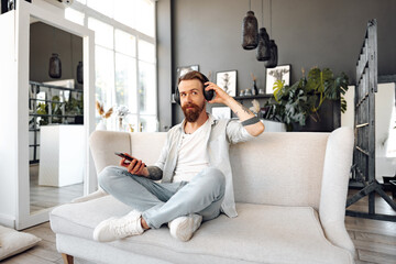 Positive bearded man listening music with headphones while sitting on couch at home