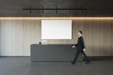 Poster - Side view of young businessman walking in modern concrete and wooden office reception interior with empty white mock up banner and desk. Lobby concept. Worker and CEO concept.