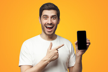 Wall Mural - Excited screaming WOW man in t-shirt presenting phone pointing at blank screen on yellow background